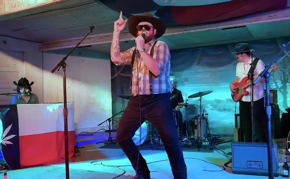 A band performs on stage, with a singer in a hat and plaid shirt in the foreground.