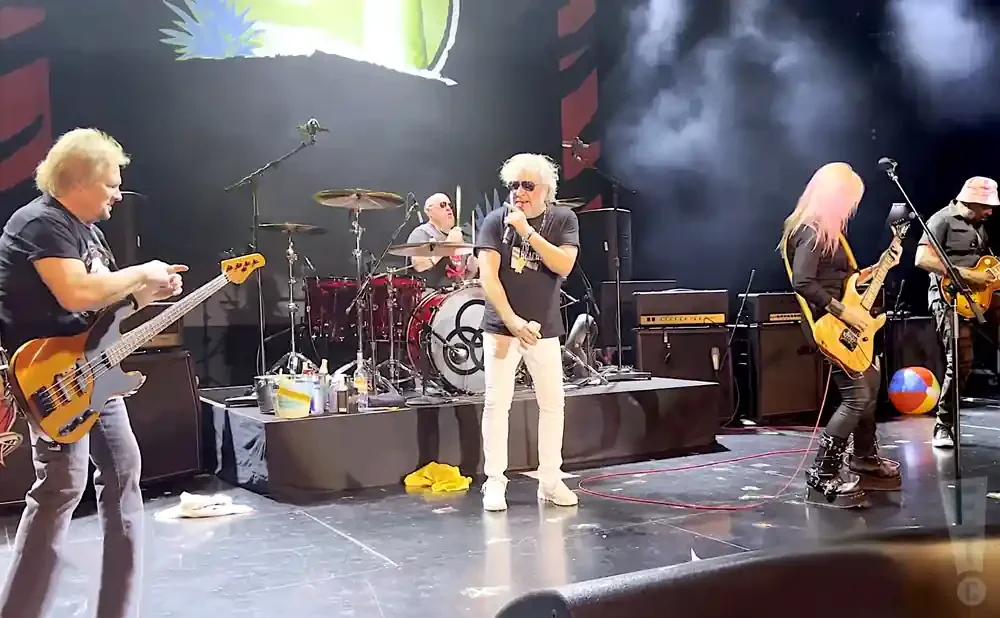 A rock band performs on stage, with a bassist on the left, a singer center, a drummer behind, and two guitarists on the right.