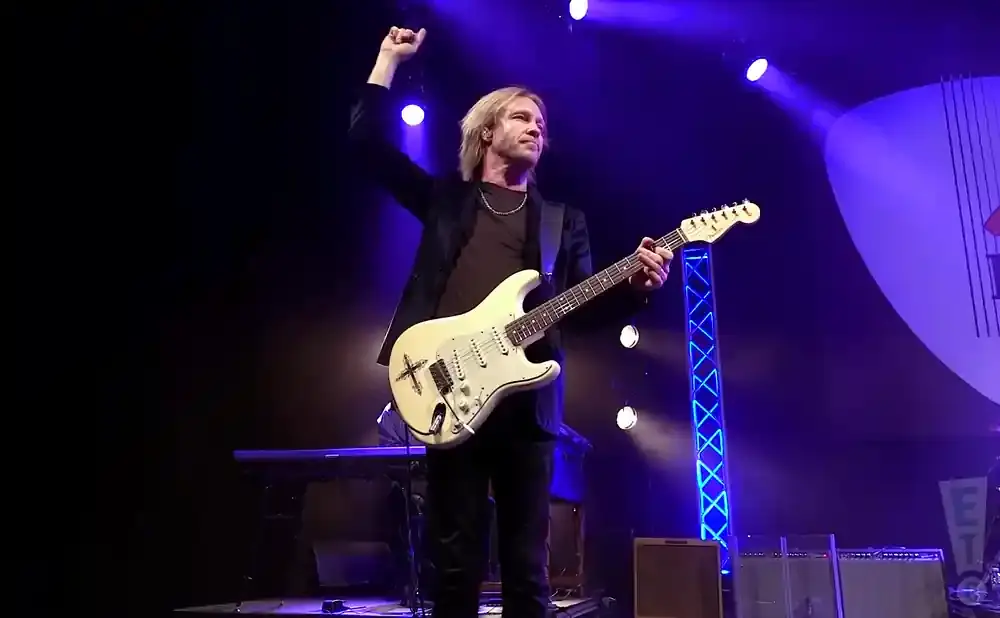 A male guitarist with blonde hair plays a white Fender Stratocaster on a dimly lit stage, lit by blue spotlights.