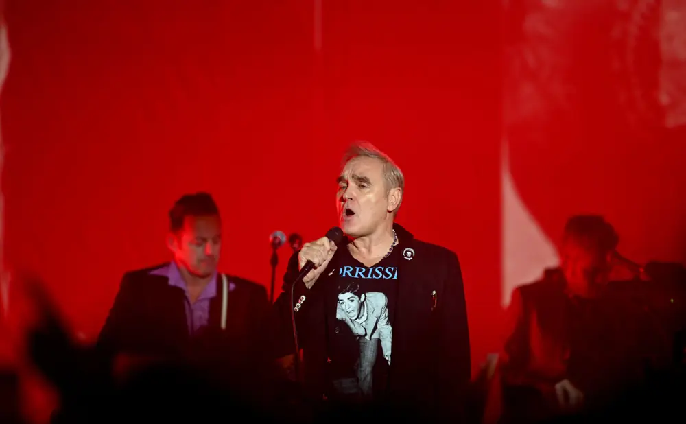 Singer Morrissey on stage in front of a red backdrop, wearing a black jacket over a t-shirt with his face on it.