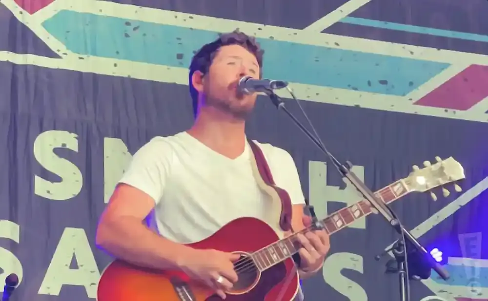 A man in a white t-shirt plays a red acoustic guitar on stage, singing into a microphone.