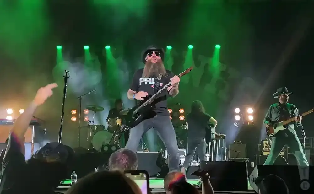 A rock band performs on a stage with green lights and smoke. The lead guitarist has a long beard and plays a black guitar.