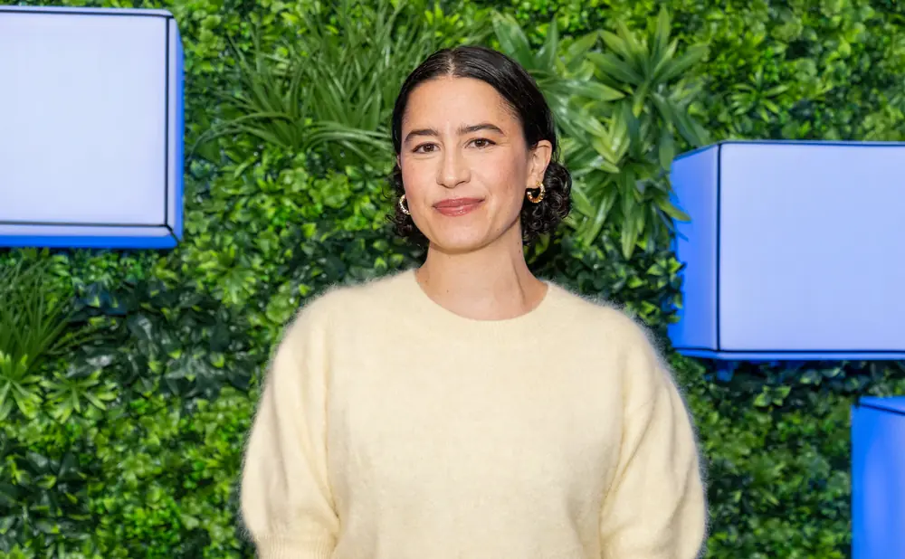 Woman in a cream sweater smiles in front of a green leafy backdrop with blue accents.