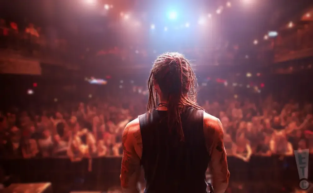 A performer with dreadlocks faces a cheering crowd on a dimly lit stage, bathed in bright spotlights.