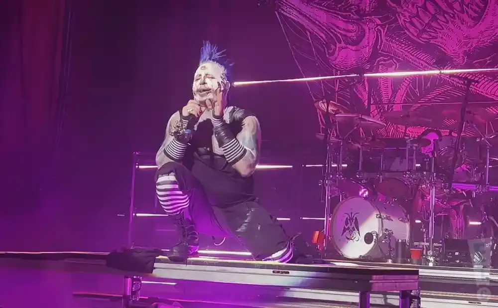 A man with blue mohawk and white face paint crouches on stage with a microphone.