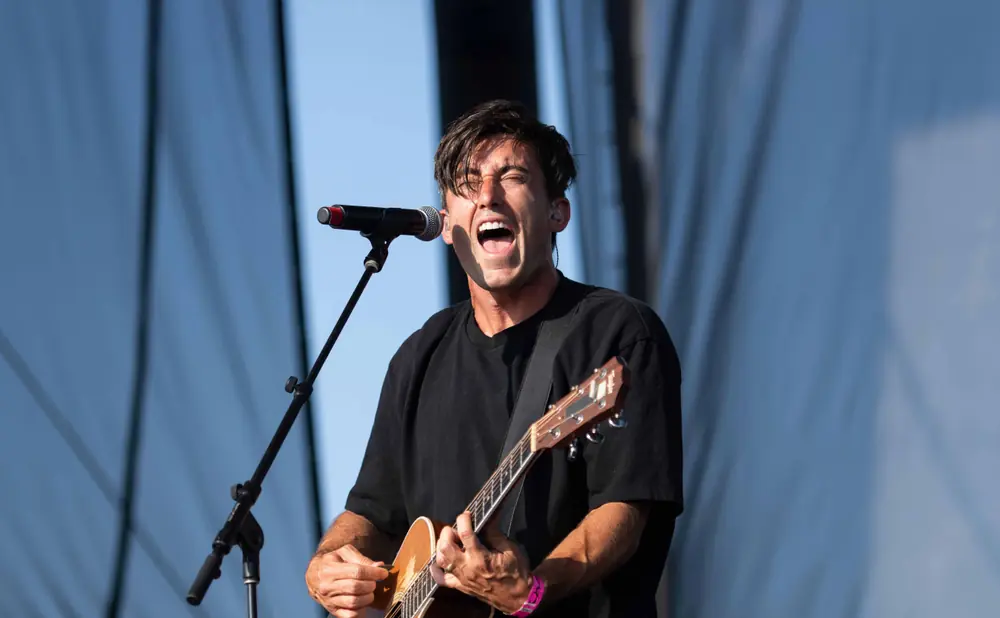 A man with dark hair sings into a microphone while playing an acoustic guitar on a stage.