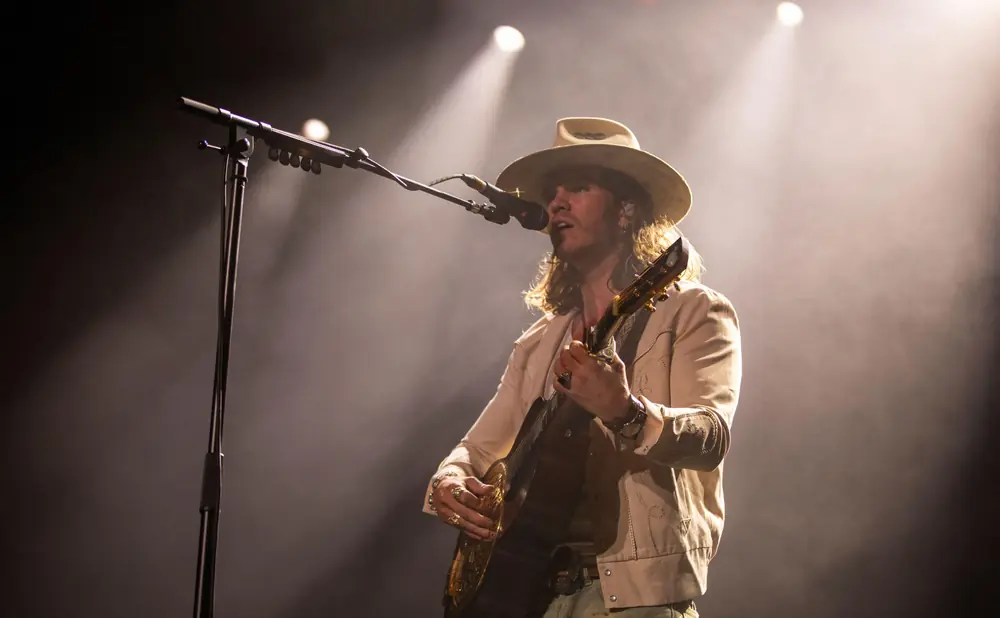 A man in a cowboy hat plays guitar on stage with spotlights shining on him.