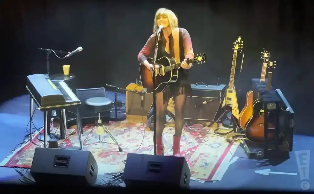 Singer plays acoustic guitar on stage with other guitars and a keyboard.