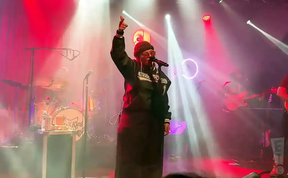 Singer in a beanie and glasses on stage, with a microphone and a band playing in the background.