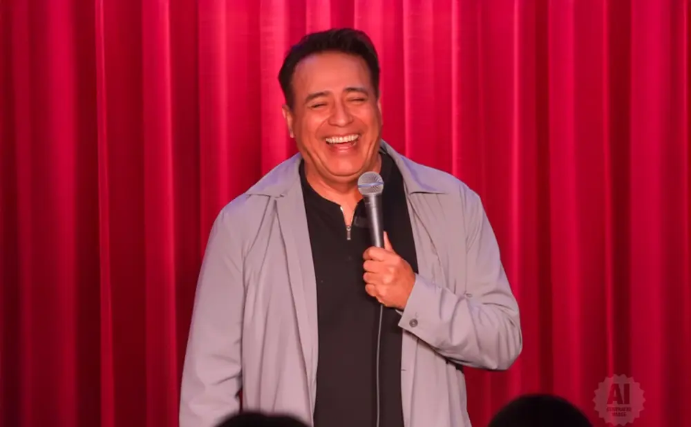 A man in a grey jacket and black shirt laughs into a microphone on a red curtain background.