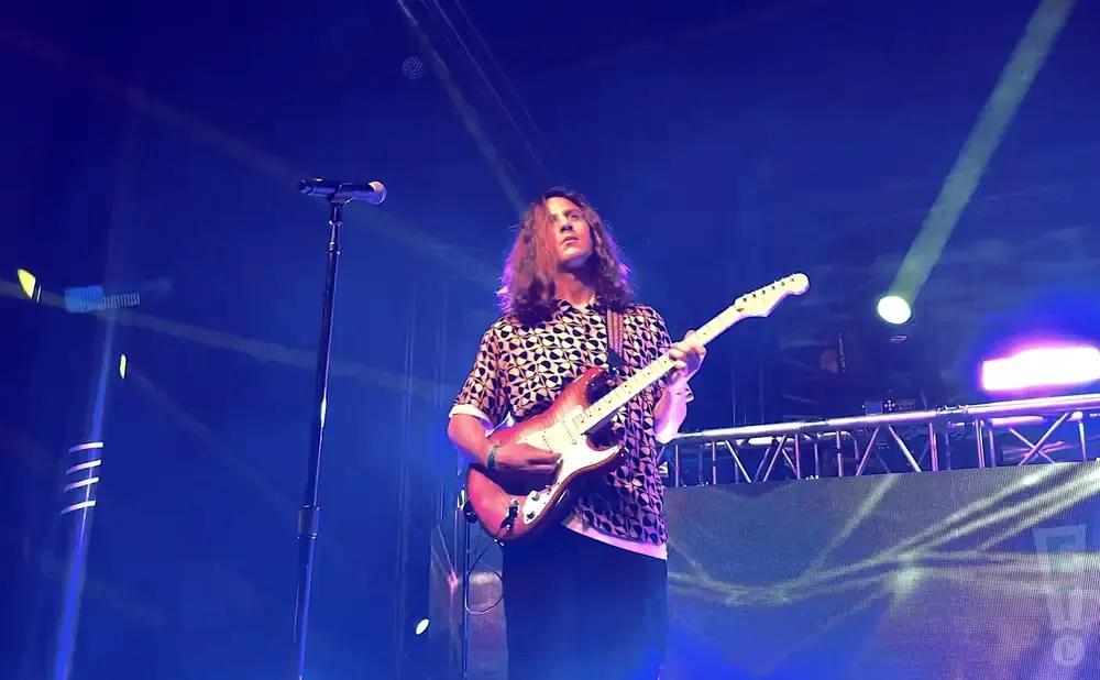 A male guitarist plays an electric guitar on a stage with blue lights.