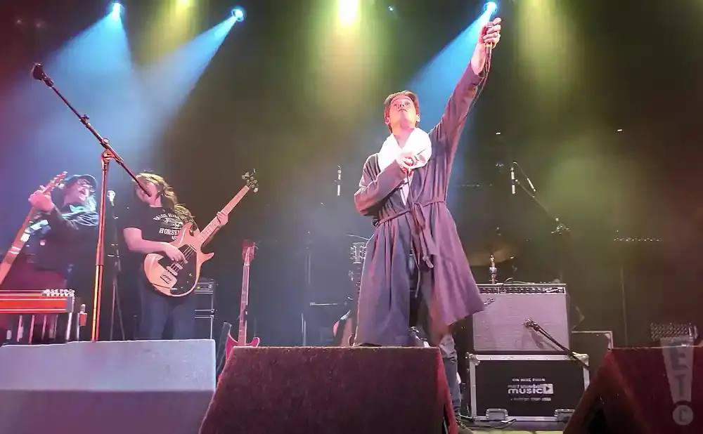 A lead singer in a robe raises a microphone and bell onstage while musicians play guitars.