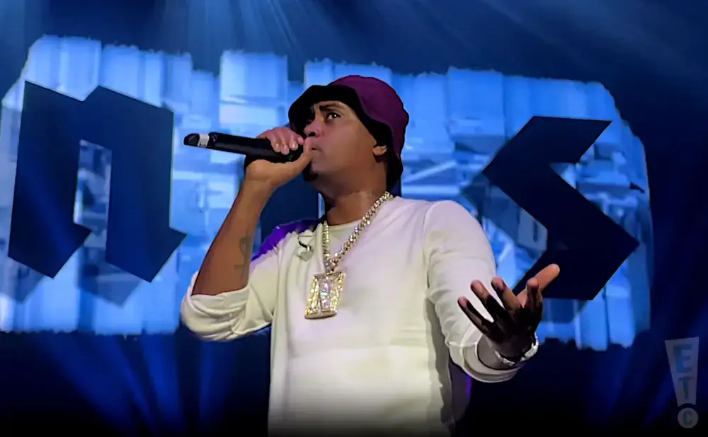 Rapper Nas wearing a white long-sleeve shirt, burgundy bucket hat, and large chain performs on stage.