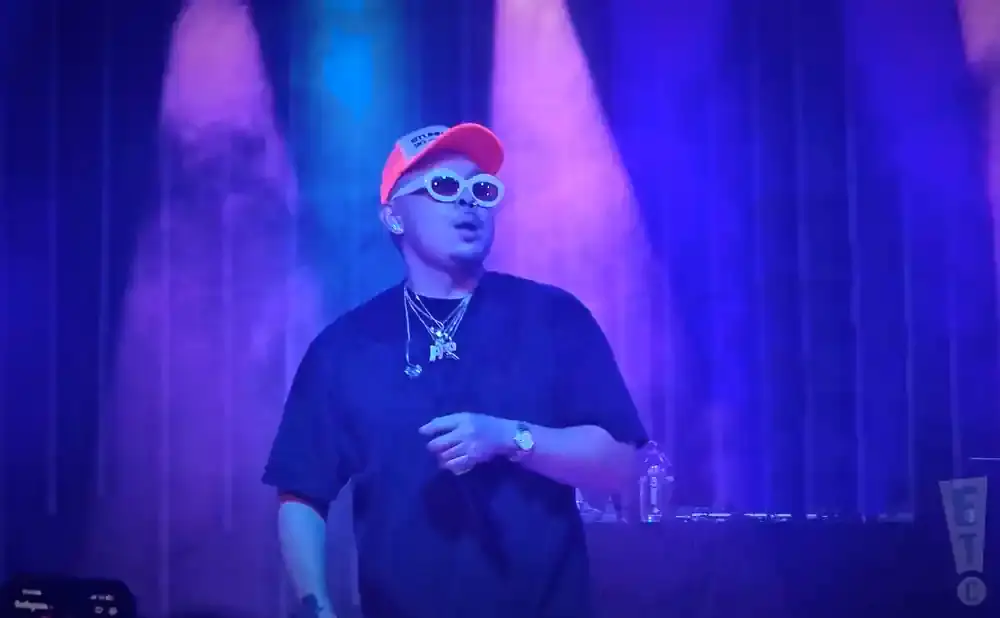 A performer in an orange hat and white oval sunglasses on a stage with purple lighting.