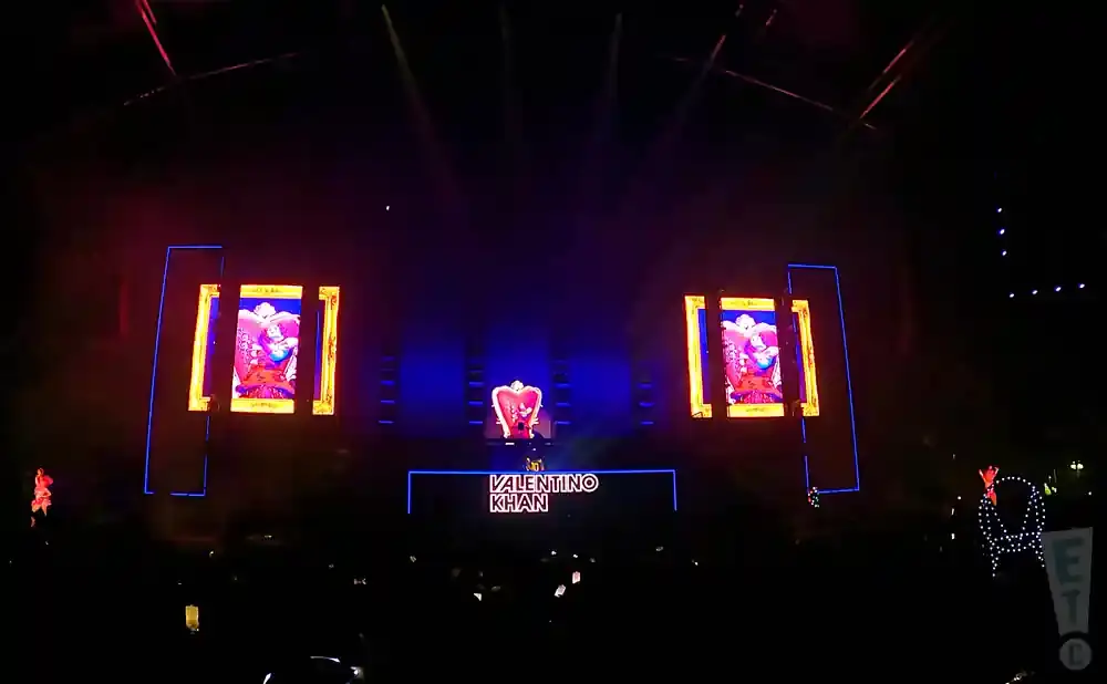 A DJ performs on a stage with large screens displaying graphics, under bright stage lights.