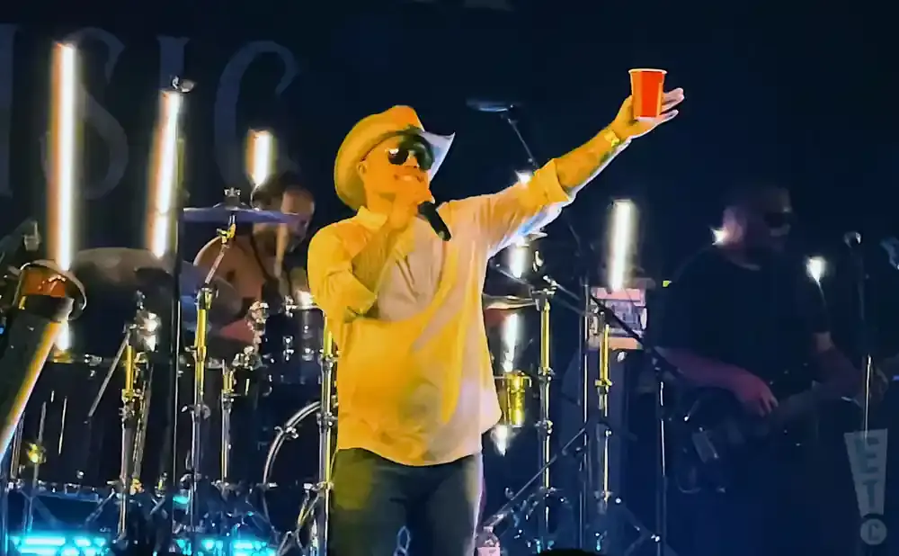 A man in a cowboy hat sings into a microphone, holding up a red cup.