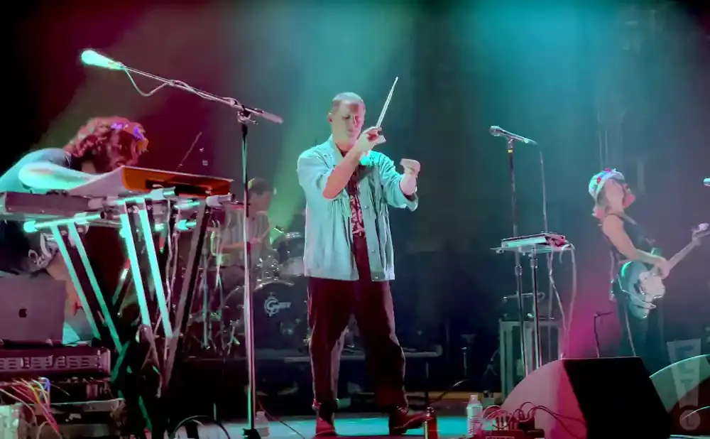 Musicians performing on stage with keyboards, a theremin, and a guitar, under colorful stage lights.
