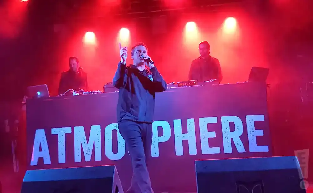 Rapper Slug from Atmosphere performs on stage with a DJ and another band member under red lights.