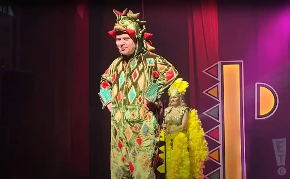 A man in a green dragon costume stands next to a woman in a yellow, feathery costume on a stage.