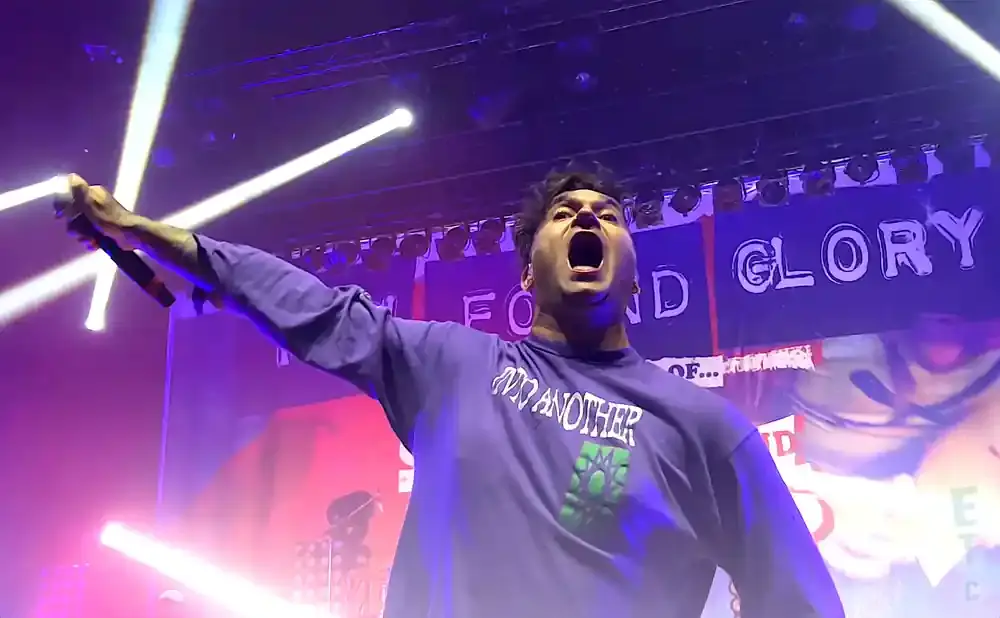 A singer in a purple shirt and green graphic yells into a microphone on a stage with bright lights.