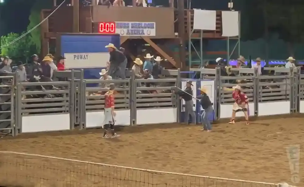 Rodeo contestants in a dirt arena, with spectators and a score board in the background.