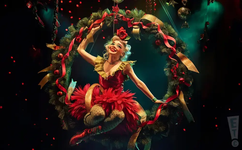 A performer in a red, feathery costume hangs from a festive Christmas wreath.
