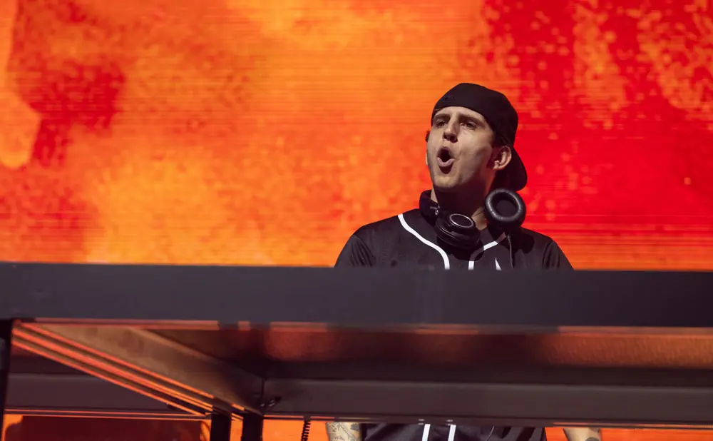 A male DJ wearing a black baseball cap and jersey with headphones around his neck performs on a stage with an orange, blurred background.
