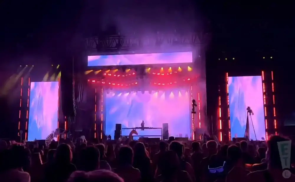 A DJ performs on stage at a concert with large screens displaying abstract visuals and a cheering crowd in the foreground.