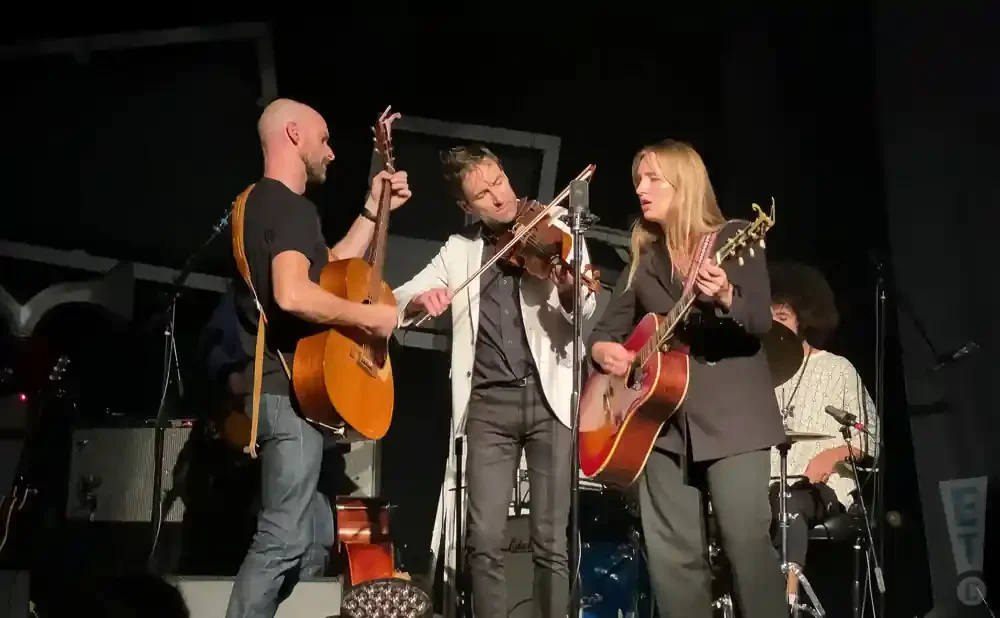 A band performs on stage, with a violinist in the center and two guitarists on either side.