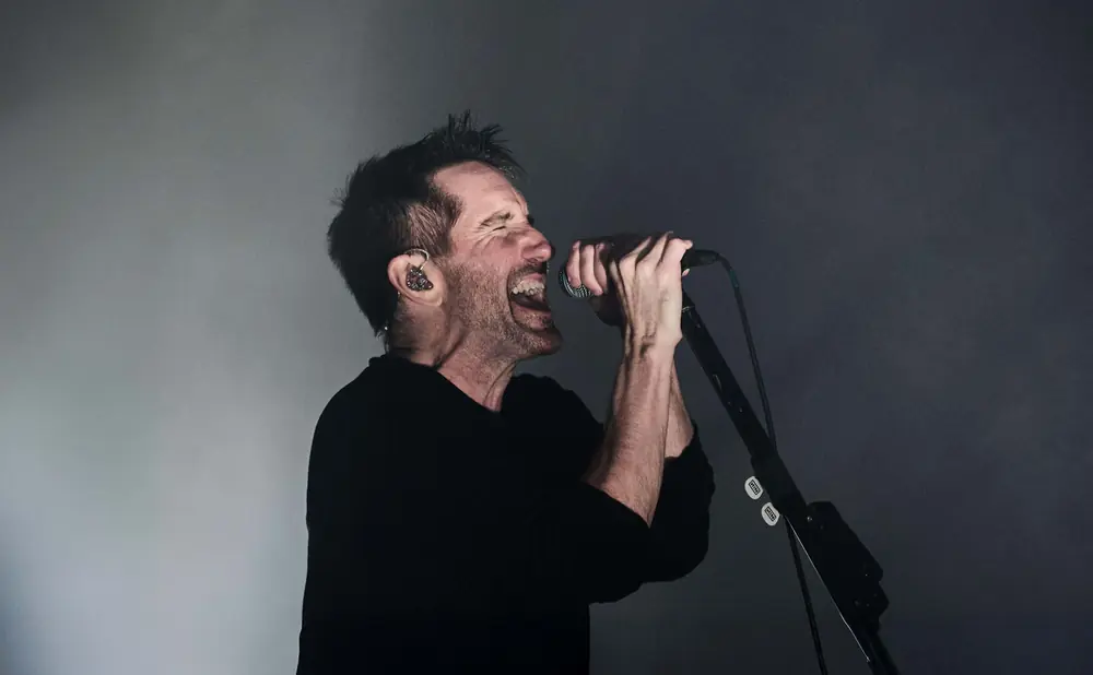 Singer with eyes closed, mouth open, gripping a microphone, wearing an earpiece, on a dark stage.