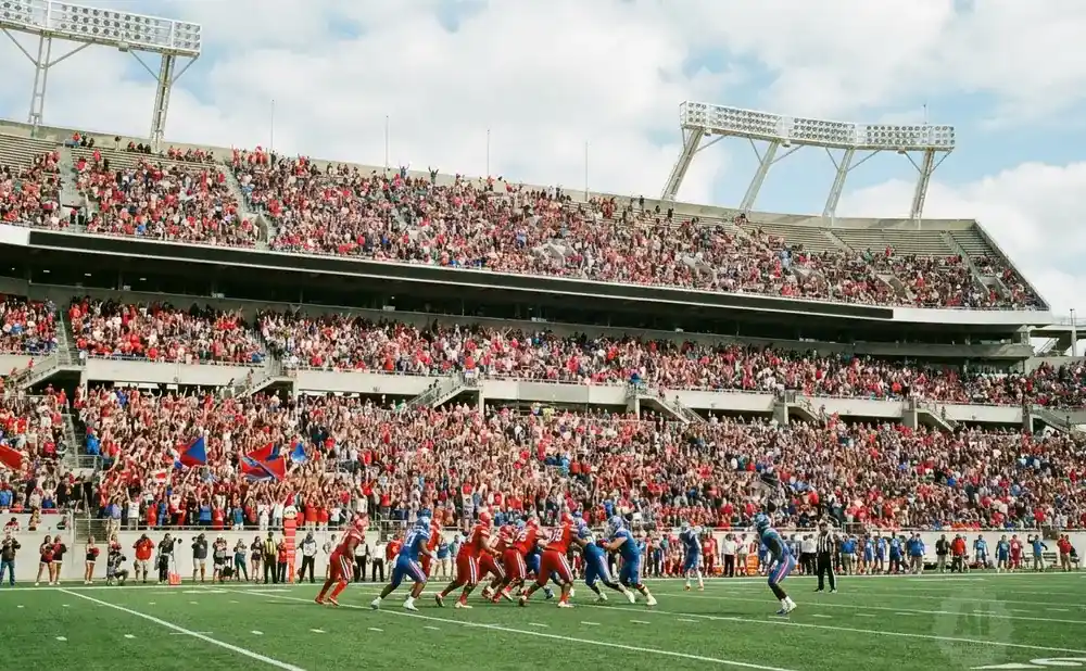 Football players in red and blue uniforms on a field, with a large crowd in the stands of a stadium under a cloudy sky.