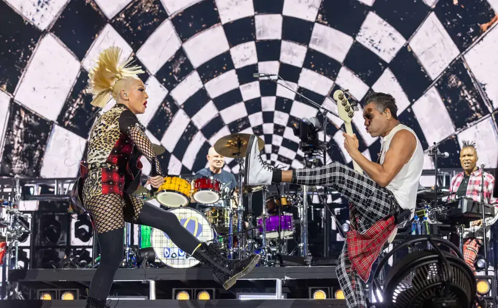 A rock band performs on stage in front of a checkered backdrop.