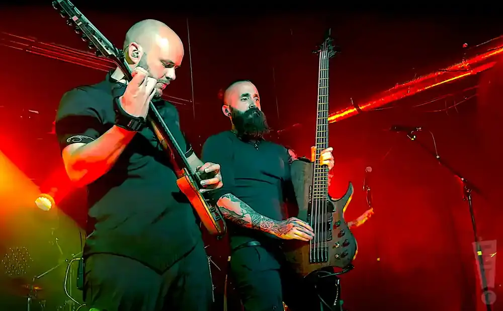 Two bald men play guitars on a stage with red lighting. One has a full beard and tattoos on his arm.