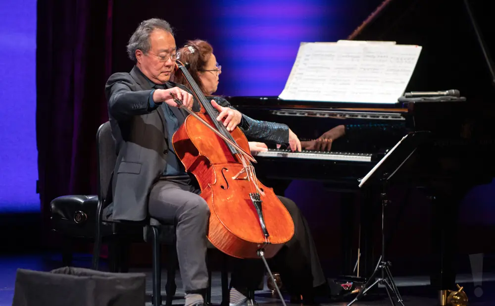 A man plays a cello next to a woman playing a piano, on a stage with purple and blue lighting.