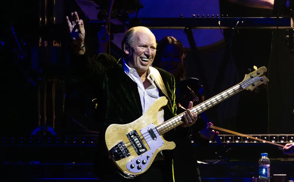 Musician playing a bass guitar on stage, waving to the audience.
