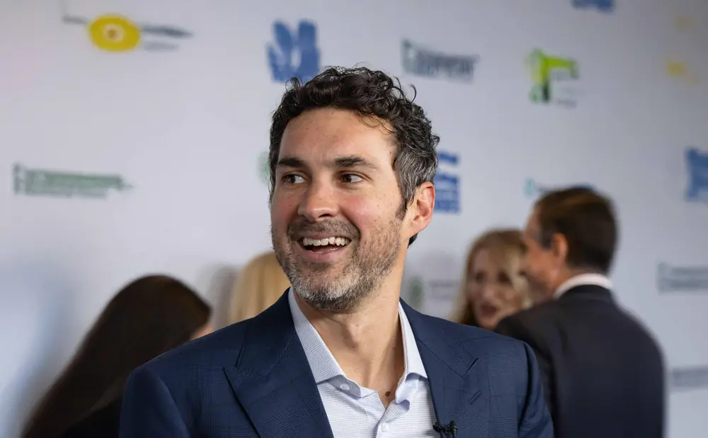 Man in a blue suit smiles at a microphone, with blurred people in the background.
