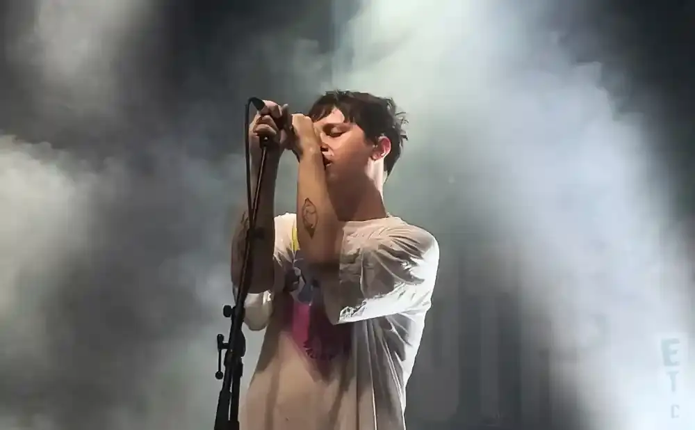 A man with dark hair sings into a microphone on a smoky stage, wearing a white t-shirt.