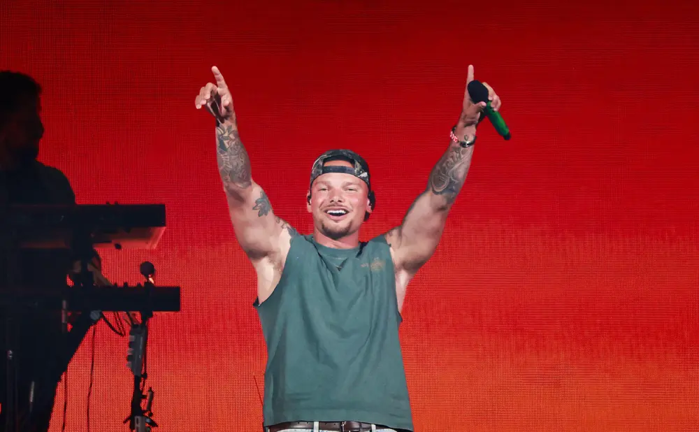 Singer Kane Brown performs on stage, wearing a green tank top, jeans, and a backward baseball cap, holding a microphone.