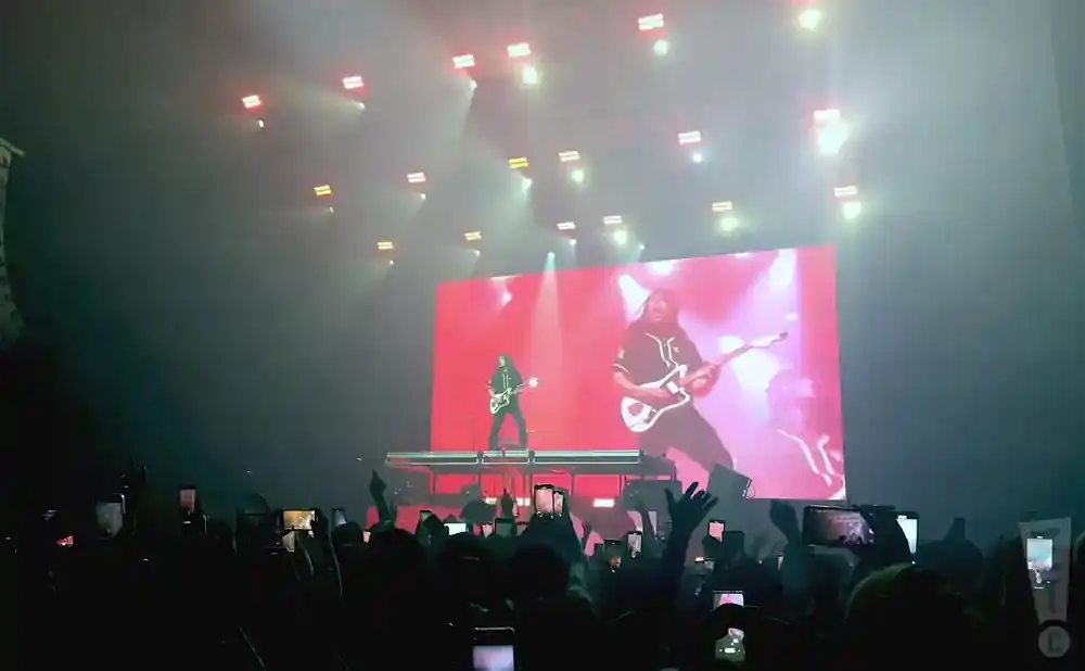 Two guitarists perform on a stage with a large red screen displaying their images, while a crowd of people hold up their phones to record.