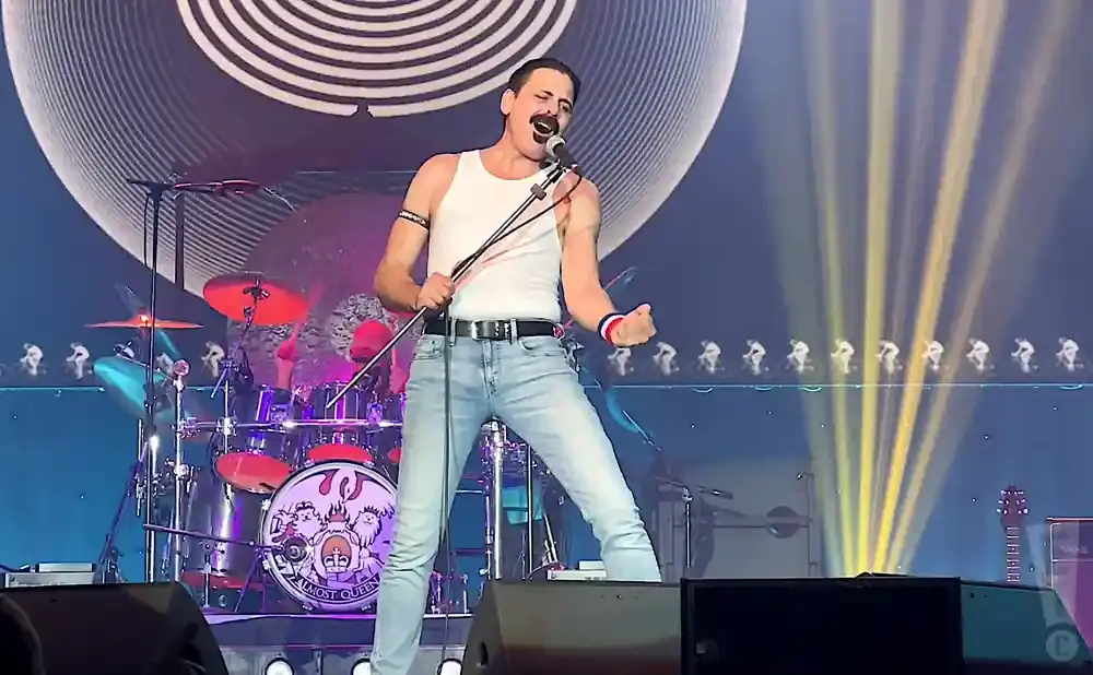 A Freddie Mercury impersonator performs on stage wearing a white tank top and jeans, with a drum set behind him.