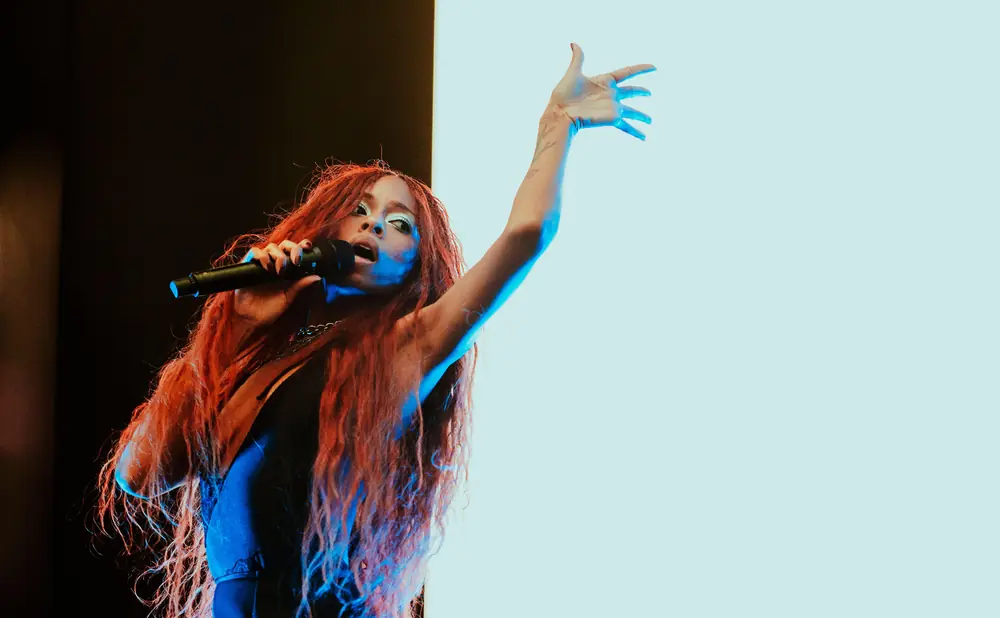 Singer with long red hair on stage, holding a microphone and reaching up with her other hand.