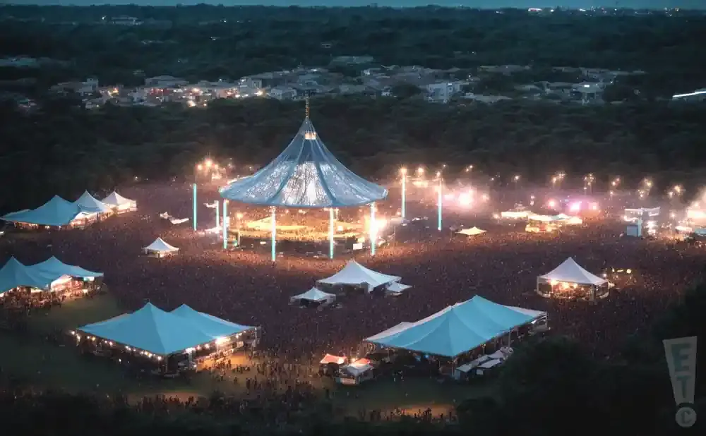An aerial view of a large outdoor music festival at dusk, with illuminated tents and a central stage structure.