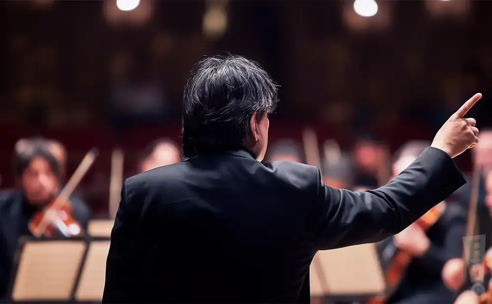 Conductor in a black suit with his back to the camera, pointing with his right hand to the right.