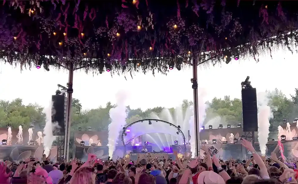 A crowd at an outdoor music festival with smoke effects and a DJ on stage.