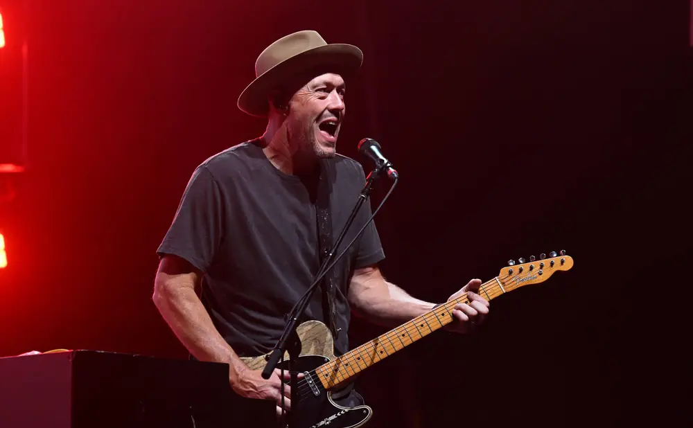 A man wearing a hat and dark t-shirt plays a guitar and sings into a microphone on a red-lit stage.
