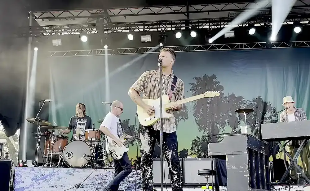 A band performs on stage with a palm tree backdrop. The lead singer plays a light-colored electric guitar.