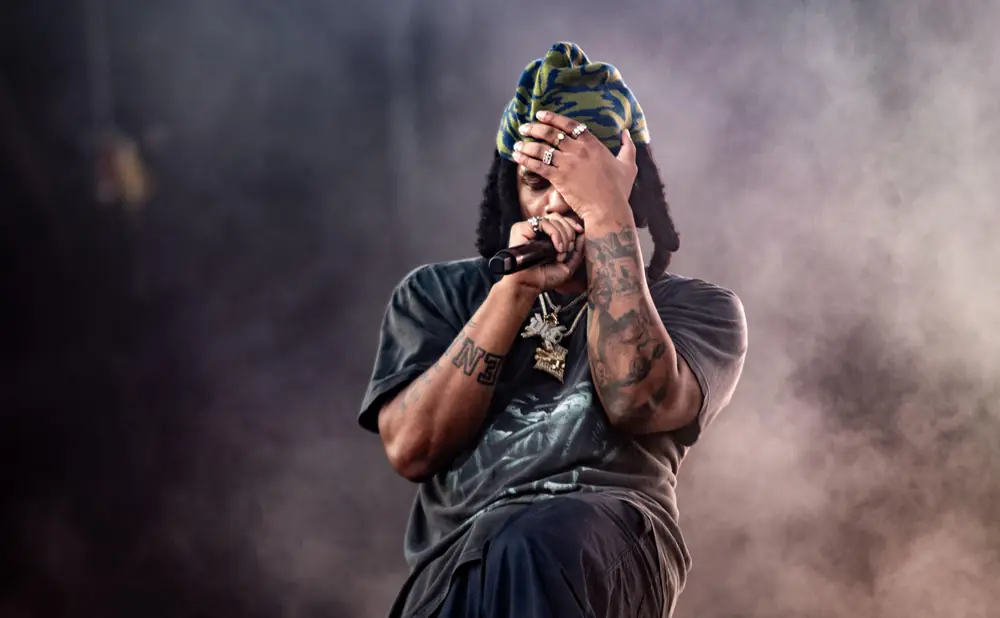 A rapper with a beanie, jewelry, and tattoos holds a microphone and covers their face.