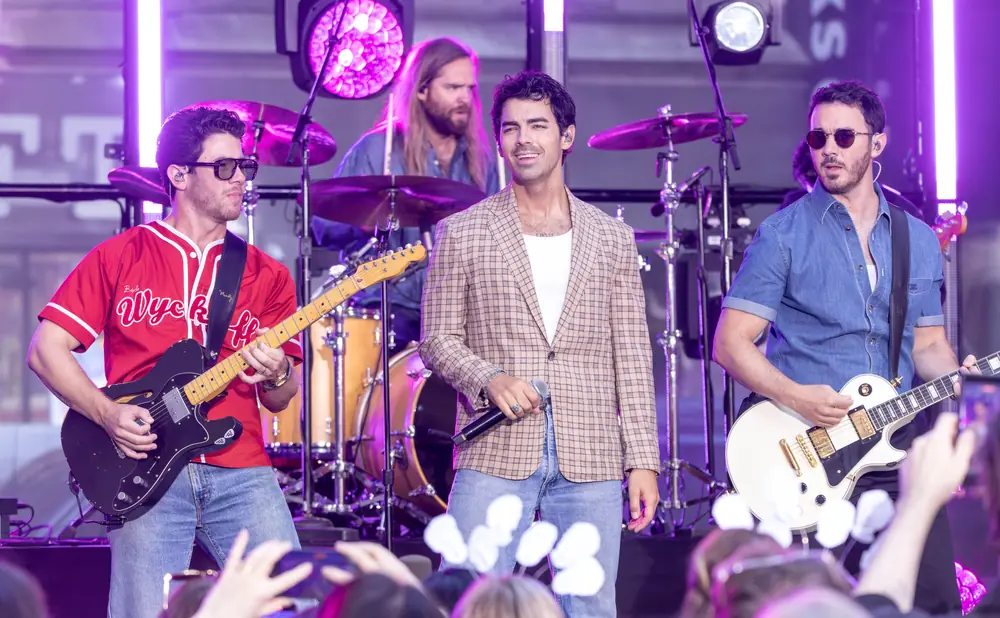 Three men on stage with instruments, one in a red baseball jersey, one in a plaid jacket, and one in a denim shirt.