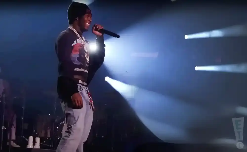 A man in a beanie, crop top, and jeans sings into a microphone on a stage with dramatic lighting.