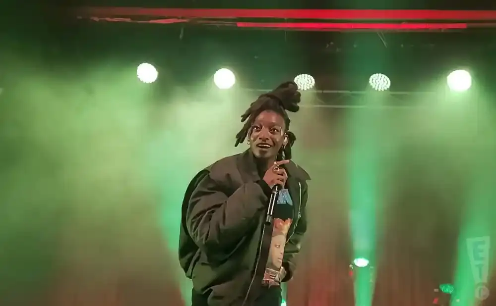 A Black person with dreadlocks sings into a microphone on a stage with green lighting.